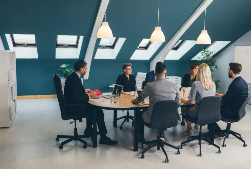 A group of business professionals in formal attire gathered around a large oval table for a meeting in a modern