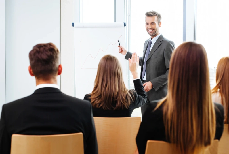 A smiling man in a grey suit and blue tie presents a data chart on a flip chart to a seated audience in a bright office.